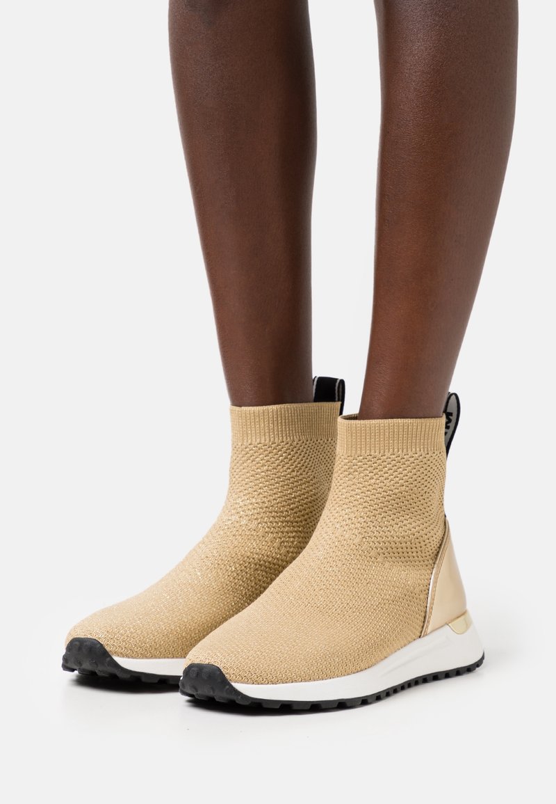 Jambes portant des baskets montantes beiges en maille style chaussette avec semelles blanches et languettes noires sur fond blanc.