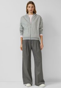 Grijze hoodie met rits en capuchon, voorzien van geribbelde manchetten. Gecombineerd met grijze wijde broek met krijtstrepen en witte sneakers.