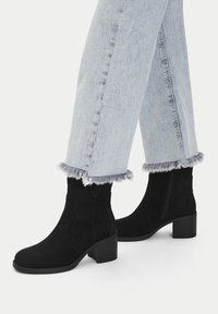 Černé semišové kotníkové boty s blokovým podpatkem, zdobené zakřivenými švy. Spárovány s světle modrými denimovými džíny s třepenými lemy.