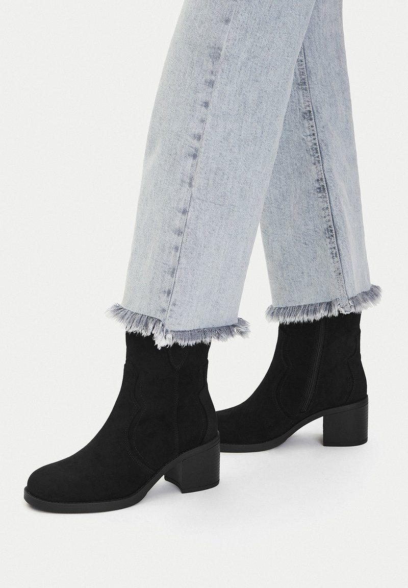 Černé semišové kotníkové boty s blokovým podpatkem, zdobené zakřivenými švy. Spárovány s světle modrými denimovými džíny s třepenými lemy.