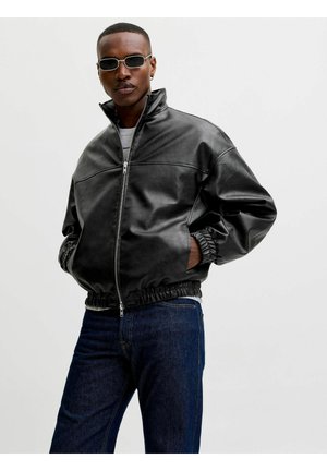 Mann iført svart skinnjakke, mørke jeans, solbriller, hender i lommer, står mot enkel lys bakgrunn.