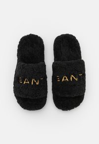 Paire de chaussons noirs duveteux à bout ouvert avec des brides rembourrées présentant un texte brodé jaune partiellement visible sur fond blanc.