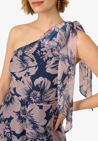 Eén-schouder bloemenjurk met een marineblauwe basis en grote roze en witte bloemenpatronen. Transparante, vloeiende stof accentueert de schouder en arm.