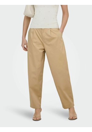 Nő bézs színű, széles szárú nadrágban, fehér, gyöngydíszítéses ujjatlan felsőben, bézs táskát tart, és áttetsző pántos, magassarkú szandált visel.