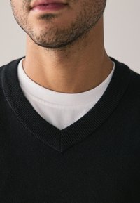 Fekete V-nyakú pulóver bordázott nyakkivágással, fehér kerek nyakú póló felett rétegezve. Közeli kép a ruha szövetének textúrájáról.