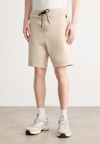 Beige šorti ar elastīgu jostu un aukliņu. Iespējas ar rāvējslēdzēja sānu kabatām un plākstera detaļu uz augšstilba. Gluda tekstūra.