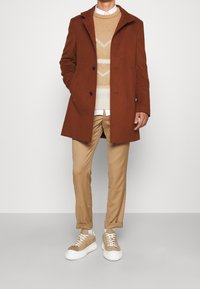 Manteau en mélange de laine marron avec un col cranté, associé à un pull crème à motifs, un pantalon beige et des baskets marron clair.