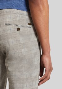 Hellgraue karierte Hose mit glatter Textur, ausgestattet mit einer einzelnen Gesäßtasche mit Knopfverschluss und einem goldenen Logo-Akzent.