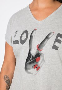 Camiseta gris de cuello en V con un gráfico en blanco y negro de una mano con uñas rojas haciendo un signo de paz, junto con la palabra "LOVE" en letras grandes.