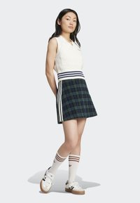 Top sem mangas de malha creme com colarinho alto, combinado com uma saia xadrez verde escura com riscas brancas. Ténis brancos e pretos com meias riscadas.