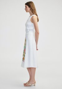 Weißes ärmelloses Kleid mit tailliertem Oberteil und schwingendem Rock, der auf einer Seite mit buntem floralem Stickerei verziert ist. Trägt beiges High-Heel-Sandalen.
