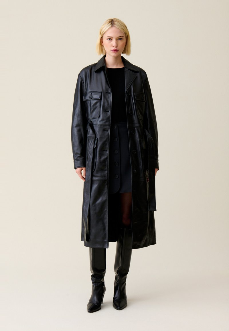 Jeune femme aux cheveux blonds courts portant un long manteau en cuir noir, une jupe noire, un haut noir et des bottes noires montant jusqu'aux genoux sur un fond beige.