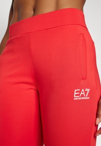 Červené športové legíny so hladkou textúrou, s bočnou vreckom a bielym logom EA7 Emporio Armani na dolnej časti nohy.