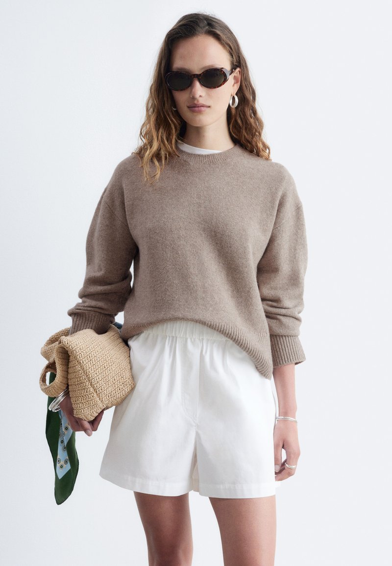 Femme portant un pull marron, un short blanc et des lunettes de soleil foncées tenant un sac tricoté beige avec une écharpe verte et bleue sur fond blanc.