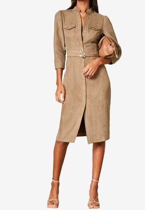 Mujer vistiendo un vestido marrón claro con cinturón hasta la rodilla y bolsillos delanteros, sosteniendo un bolso de mano a juego y llevando sandalias de tacón alto con tiras.