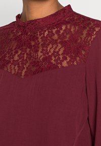 Top bordeaux con un collo alto in pizzo e motivi floreali, con un tessuto inferiore liscio e maniche lunghe.