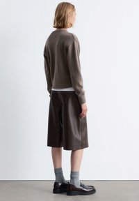 Brauner Strickpullover mit rundem Halsausschnitt, braune Ledershorts, graue Socken und schwarze, glänzende Slipper, von hinten betrachtet.