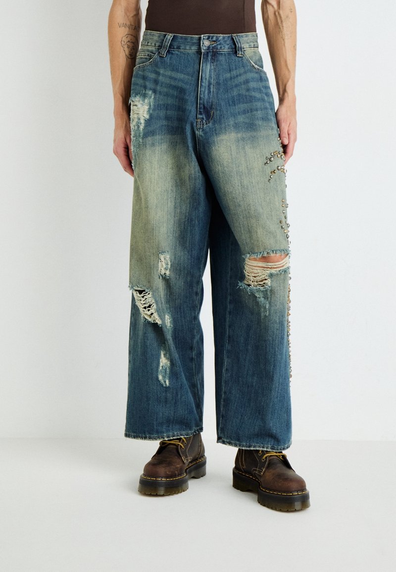 Jeans de mezclilla azul de pierna ancha con detalles desgastados, grandes rasgaduras y acentos de tachuelas plateadas a lo largo del costado. Combinados con botas marrones con cordones.