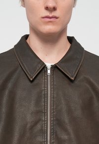 Chaqueta de cuero marrón con un acabado texturizado, que cuenta con un cierre frontal de cremallera y un cuello con solapa. El diseño es minimalista y moderno.