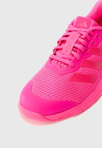 Lyse rosa sportssko med mesh-overdel, polstret tunge, justerbar snøring og teksturert såle. Har tre markante tonale striper.