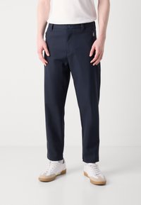 Ναυτικό μπλε παντελόνι με ίσια γραμμή, κατασκευασμένο από απαλό ύφασμα. Διαθέτει κουμπί κλεισίματος και πλαϊνές τσέπες. Συνδυασμένο με λευκά αθλητικά παπούτσια.