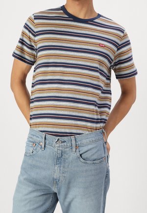Homme portant un t-shirt rayé à manches courtes avec des lignes bleues, marron et blanches et un jean en denim bleu clair, mains dans les poches.