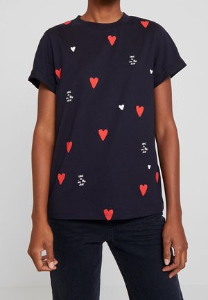 Persoon draagt een marineblauw t-shirt met rode en witte harten en witte tekst met de woorden "Love is in the Air" verspreid over de stof.