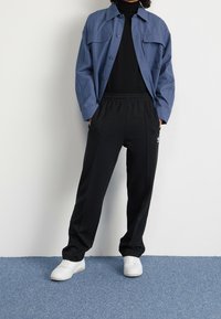 Blå skjorta med knappar och två bröstfickor, svart polotröja under, svarta mjukisbyxor och vita sneakers på en ljusblå yta.