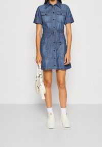 Jeansskjolklänning med korta ärmar, skjortkrage, två bröstfickor och ett knytbälte i midjan. Kombineras med en vit axelremsväska och sneakers.