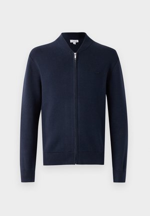 Marineblauer Strickcardigan mit Reißverschluss, Stehkragen und gerippten Bündchen. Verfügt über ein kleines Logo auf der Brust als Detail.