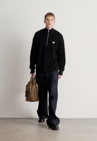 Čierny pletený sveter na zips, tmavomodré džínsy a čierne topánky. Modelka nesie hnedú kabelku cez rameno, stojí na svetlosivom pozadí.