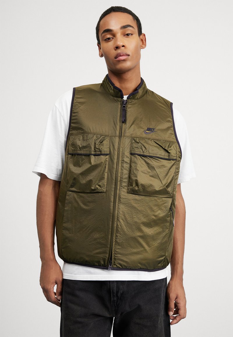 Gilet sans manches vert olive en matériau léger et brillant. Il dispose de deux poches poitrine, d'une fermeture éclair et d'un col haut. Accents noirs visibles.