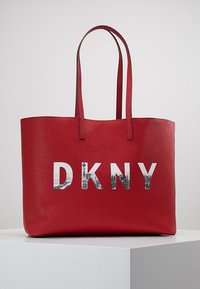 Röd tote-väska i strukturerat syntetmaterial, med dubbla handtag och en reflekterande "DKNY"-logga i vitt och grått mönster.