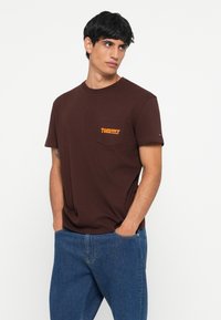 Tricou din bumbac maro, cu guler rotund și mâneci scurte. Prezintă un logo portocaliu "TOMMY" pe buzunarul din stânga pieptului. Purtați-l cu blugi albaștri.