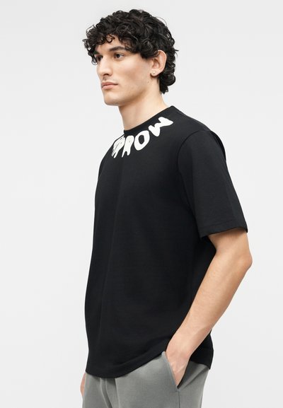 Νεαρός άνδρας με σγουρά μαλλιά, που φοράει μαύρο μπλουζάκι με λευκή επιγραφή "GROW" κοντά στο λαιμό και γκρι παντελόνι, με το χέρι στην τσέπη.