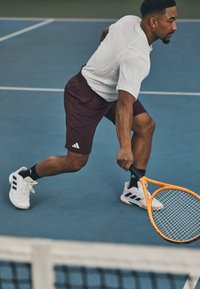 Joueur de tennis portant un maillot blanc évacuant l'humidité et un short bordeaux, avec une raquette orange, et des chaussures noires et blanches sur un court bleu.