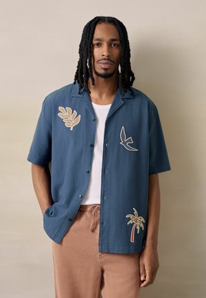 Man with shoulder-length dreadlocks wearing a blue short-sleeve shirt with tropical patches and beige pants, standing against a plain wall.