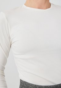 Langermet hvit topp laget av mykt, stretch-stoff med rund hals, tettsittende design og glatt tekstur.