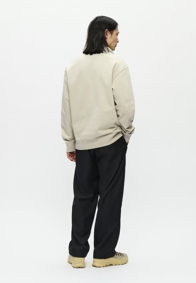 Uomo con capelli neri lunghi fino alle spalle che sta di fronte a destra, indossa un maglione beige, pantaloni neri e scarpe da trekking beige su sfondo bianco.