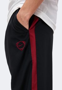 Personne portant un short de sport noir avec une bande latérale rouge et un logo Nike rouge, main reposant dans la poche, haut en mesh blanc visible.