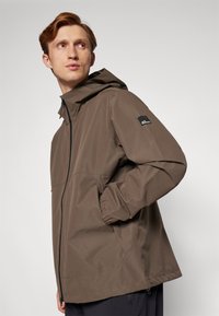 Brun vandtæt jakke med hætte, prydet med en frontlynlås, elastiske manchetter og et minimalistisk design. Sidelommer og logo-patch.