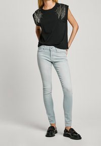 Žena v černém topu bez rukávů se stříbrnými ozdobami, světle modrých skinny džínách a černých mokasínech, stojící s rukama za zády.