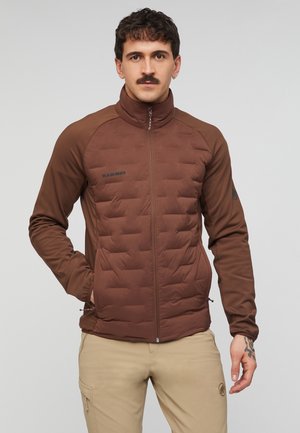 Mand med overskæg iført en brun lynlåsjakke og beige bukser, stående med den ene hånd i lommen mod en ensfarvet baggrund.