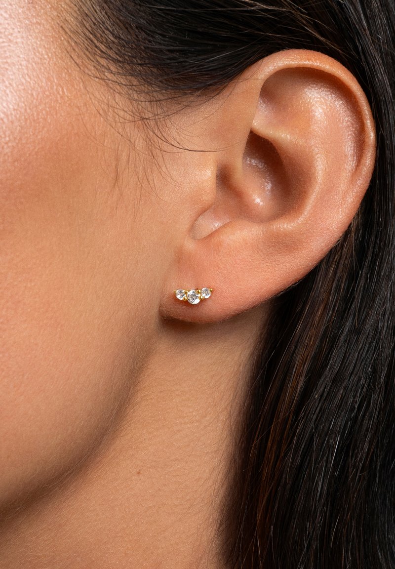 Pendientes en tono dorado que presentan tres piedras preciosas transparentes en una disposición curvada, sujetos a la oreja de un modelo. Textura de piel suave en el fondo.