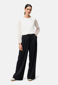 Weiße Bluse mit transparenten langen Ärmeln und vertikalen Falten, kombiniert mit hoch taillierten, weit geschnittenen schwarzen Hosen und weißen Slippern.