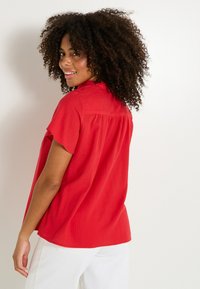Blouse rouge à manches courtes avec une coupe décontractée et un pli dans le dos. Le tissu semble léger avec une texture lisse.