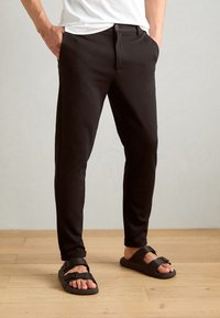 Black tailored pants with a button closure, slim fit, side pockets, and rolled cuffs, paired with black textured sandals.