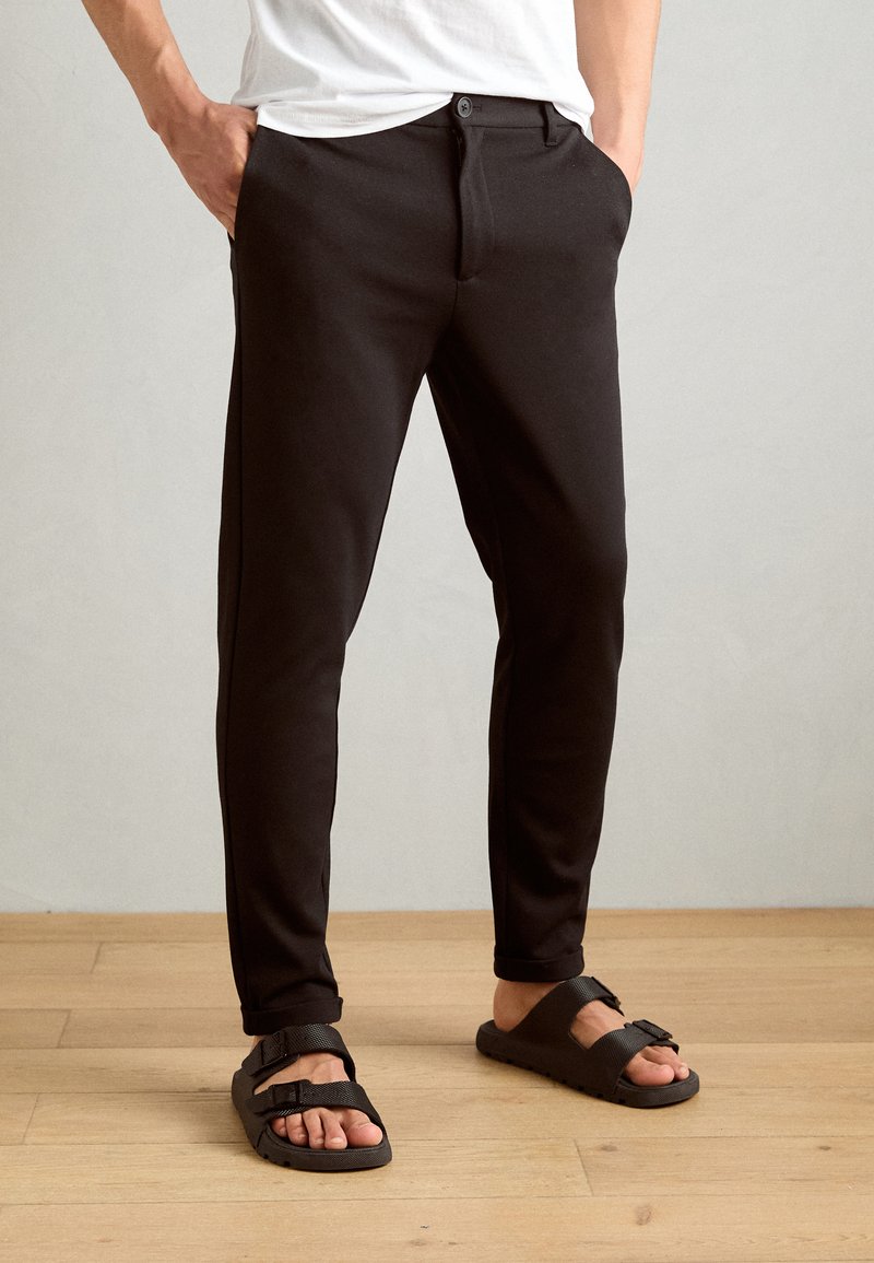 Black tailored pants with a button closure, slim fit, side pockets, and rolled cuffs, paired with black textured sandals.
