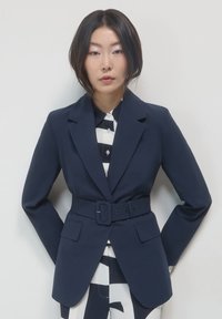 Blazer blu navy con spalle strutturate, revers, vita cintrata e due tasche frontali, indossato sopra una camicia a righe bianco e nere.