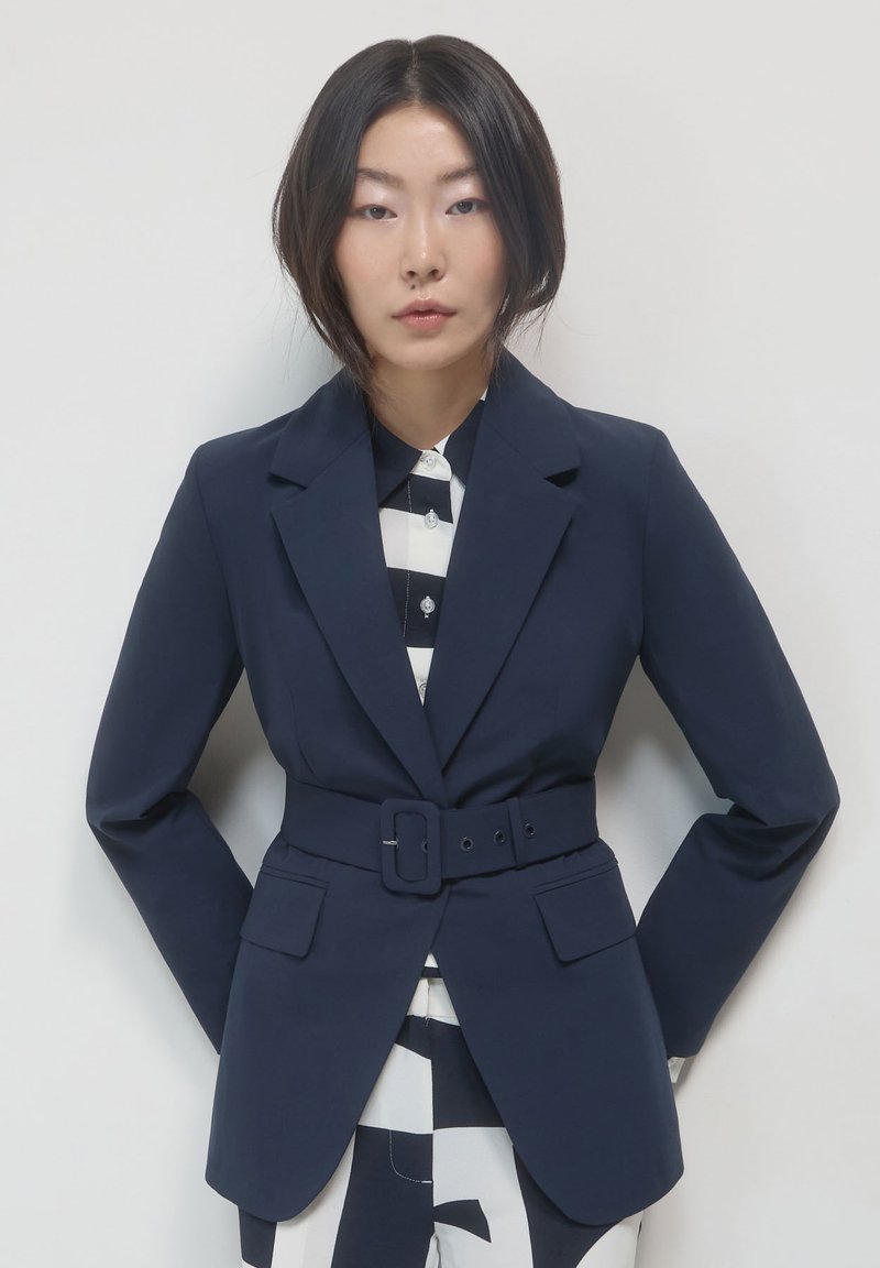 Blazer blu navy con spalle strutturate, revers, vita cintrata e due tasche frontali, indossato sopra una camicia a righe bianco e nere.